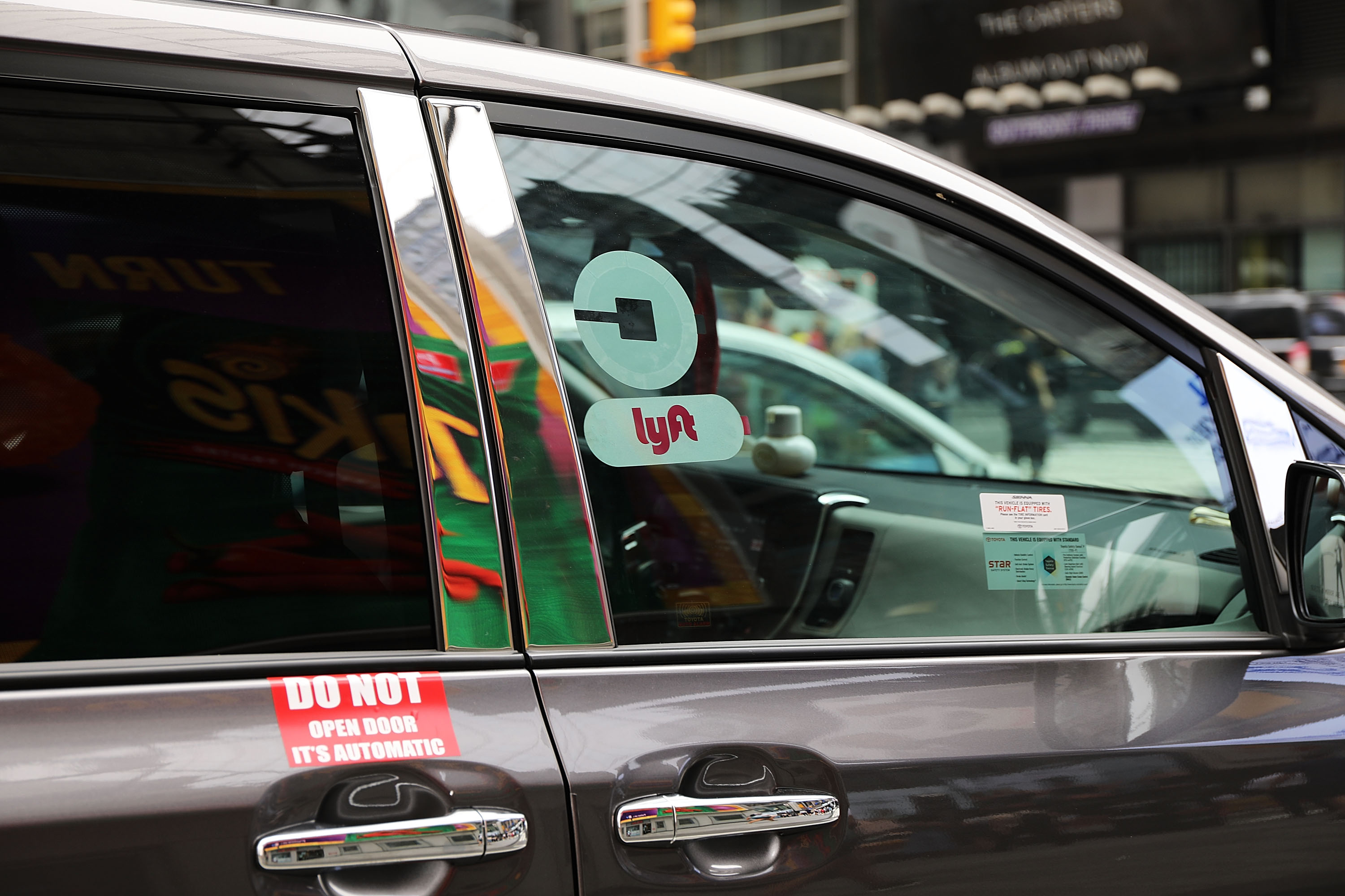 A gray car has Uber and Lyft logos on the passenger-side window