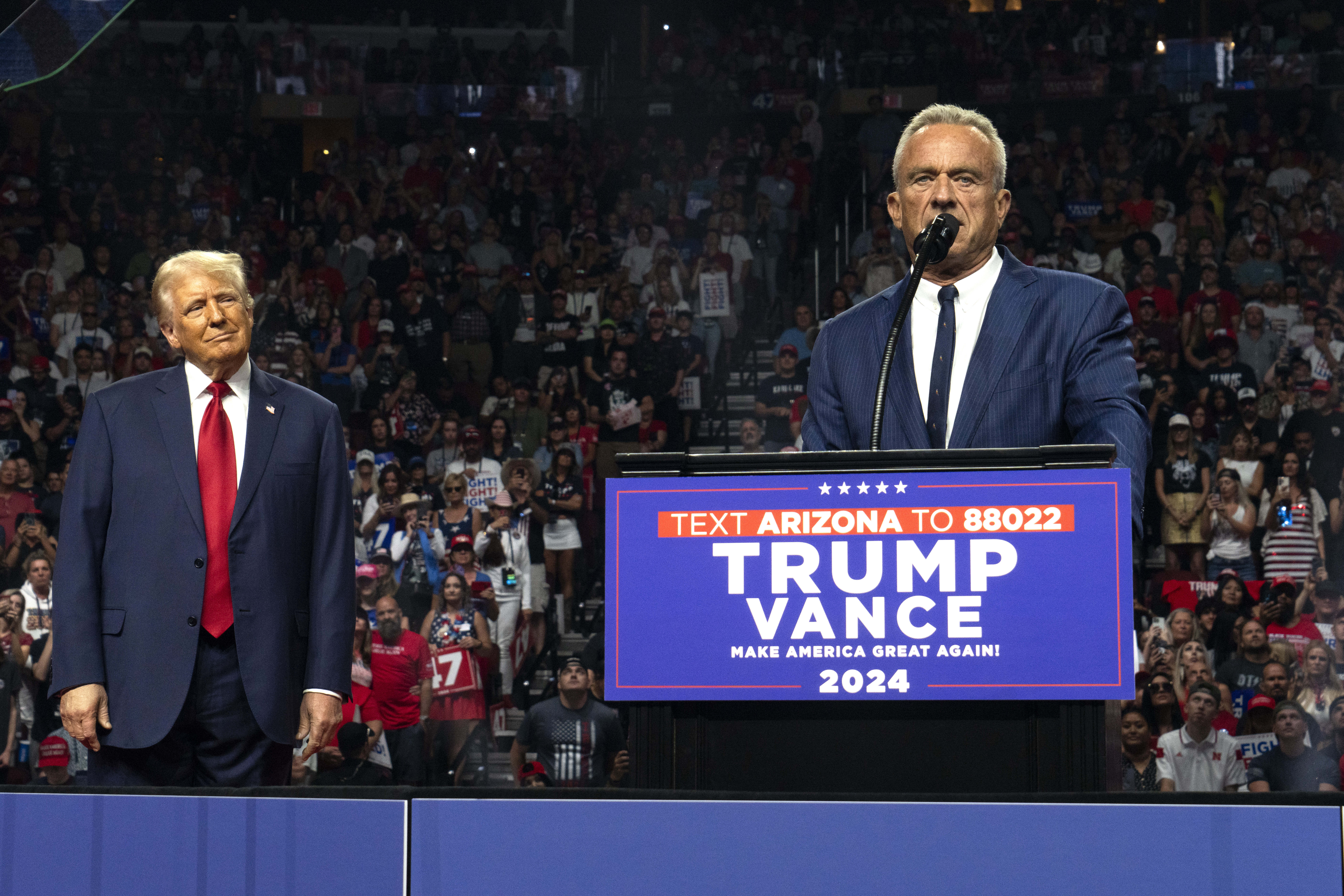 Robert F. Kennedy Jr. at the microphone of a TRUMP VANCE podium with former president Donald Trump Jr. watching from his side.