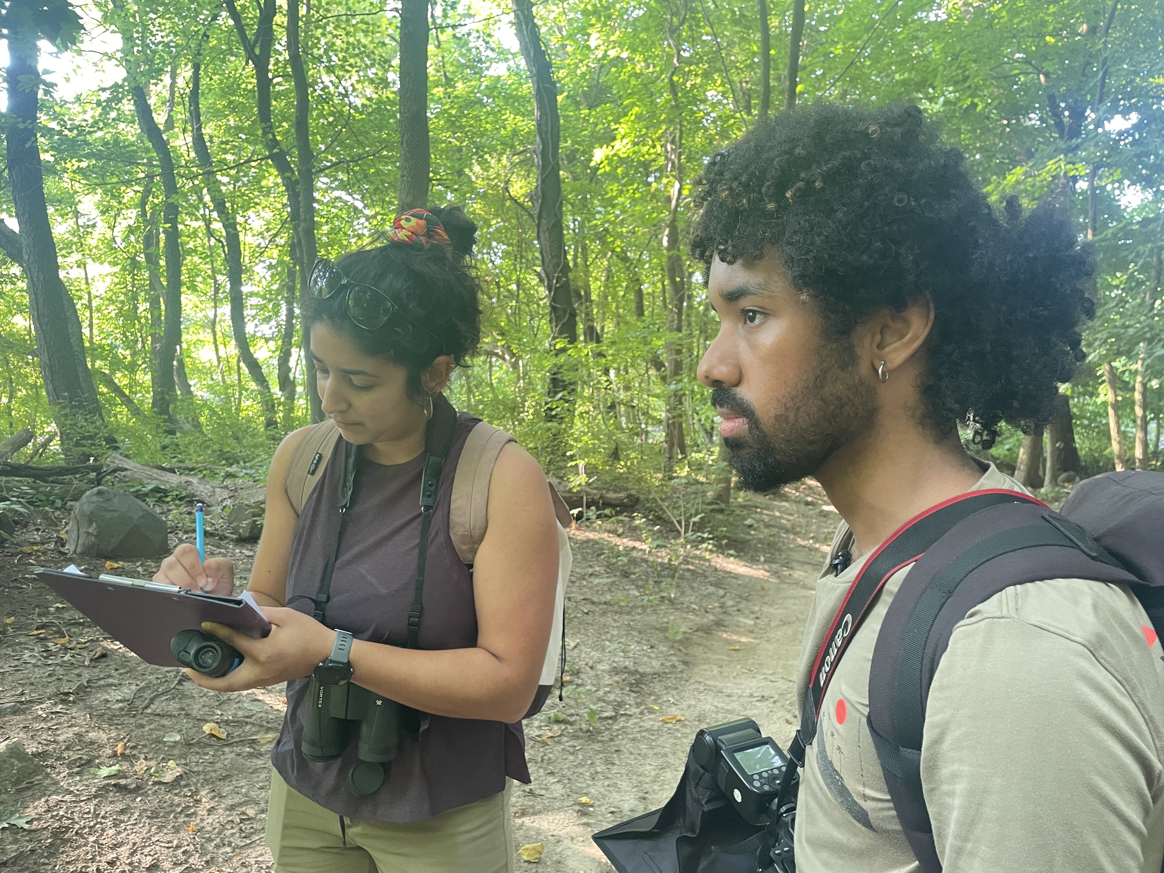A woman writes on a clipboard standing in the woods with a man.