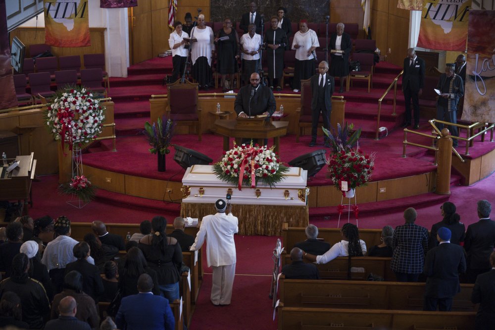 A casket sits in a carpeted sanctuary in front of a large crowd.