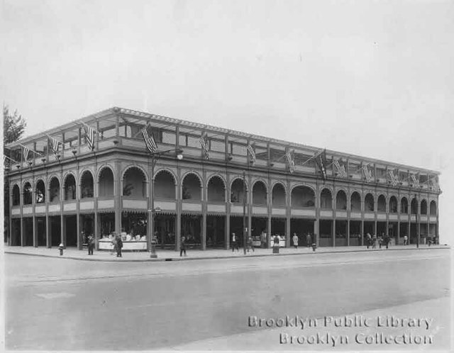 Facade of Feltman's Restaurant, a large, two-story building with roof deck on Surf Avenue at the boardwalk in Coney Island; several flags flying from roof, several pedestrians, two fire hydrants, and intersecting streets.