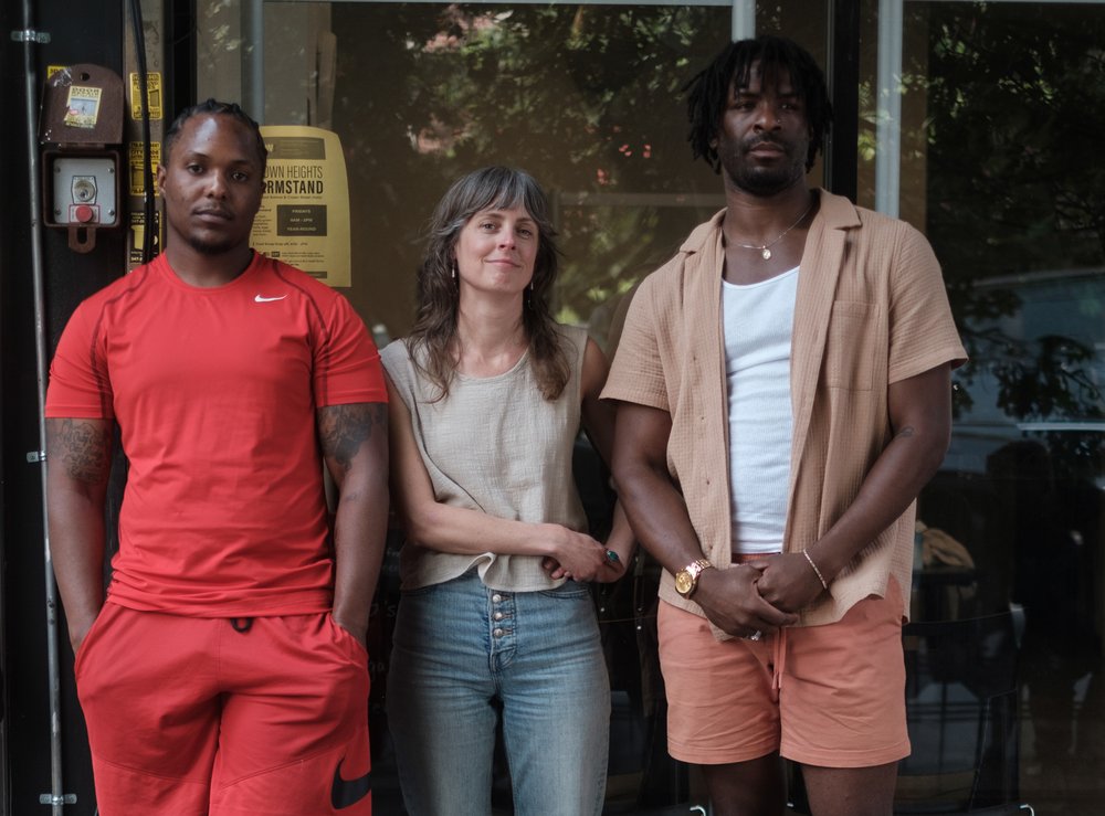 A woman wearing a plain white t-shirt stands between two men in front of the window of a store. One man is wearing a plain red shirt and red shorts and the other is wearing pink shorts, a plain white tank top and an unbuttoned tan short sleeve shirt on top.