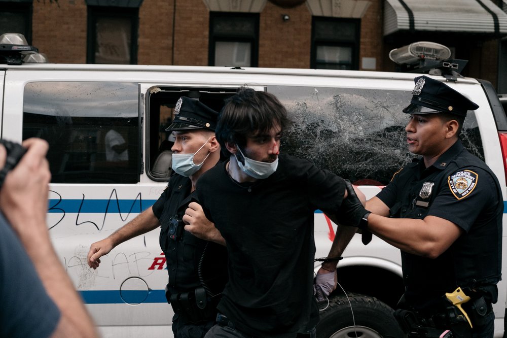 A protester with blood on his nose is led away by police, in front of an NYPD can with a cracked window