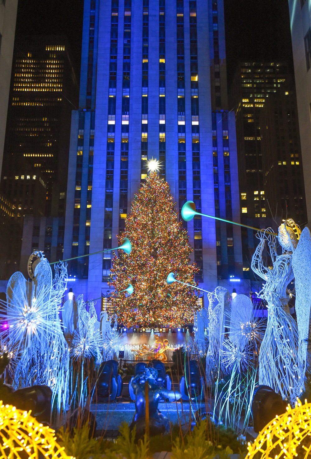 The Rockefeller Center tree is lit in multi-colored lights, within Rockefeller Center buildings