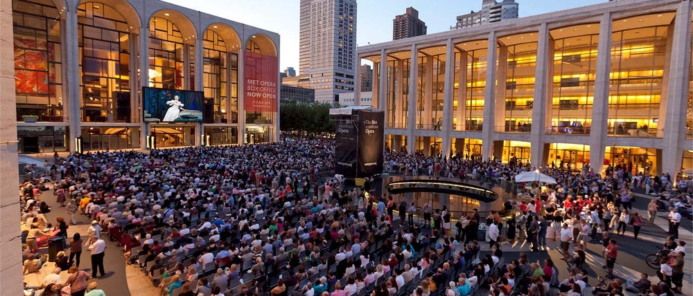 A large crowd of people seated on a plaza watching a film projected on a screen overhead.