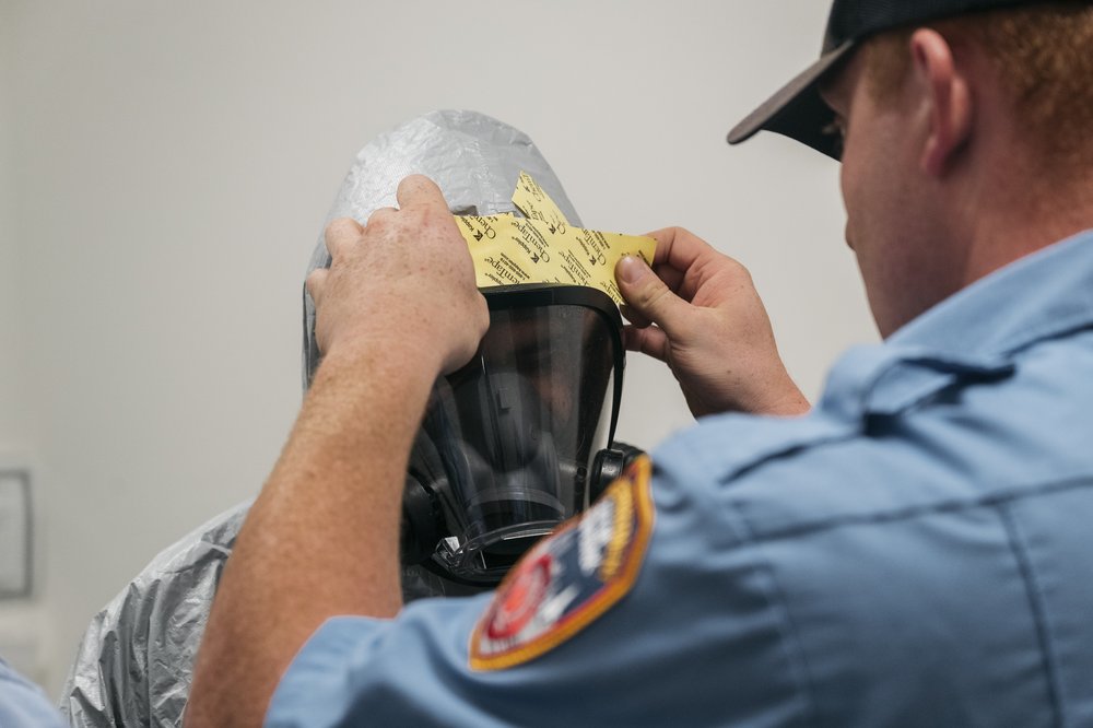 An uniformed emergency worker places an adhesive bandage-looking material on the mask of a person in full hazmat suit.