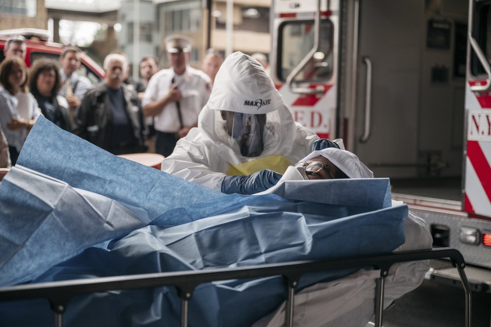 A clinician in full protective gear administers care to a person also fully clad in protective gear. The patient is laying on a stretcher.
