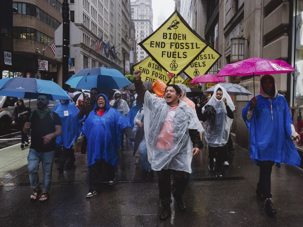 A large group of protestors, many wearing plastic ponchos, walk through rain holding signs that read: "Biden End Fossil Fuels."