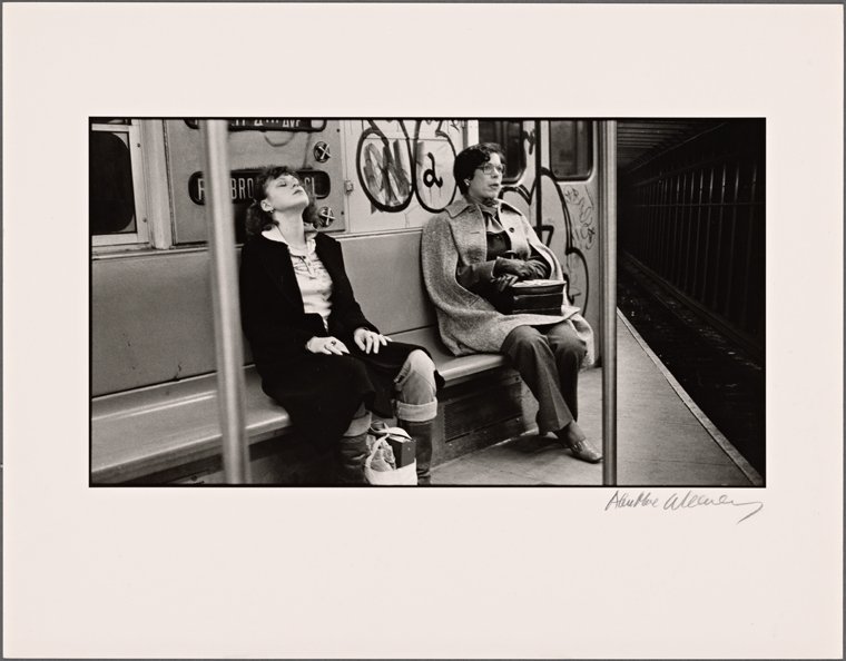 A black and white photo of two women sitting on a bench inside a subway, with a tunnel nearby.