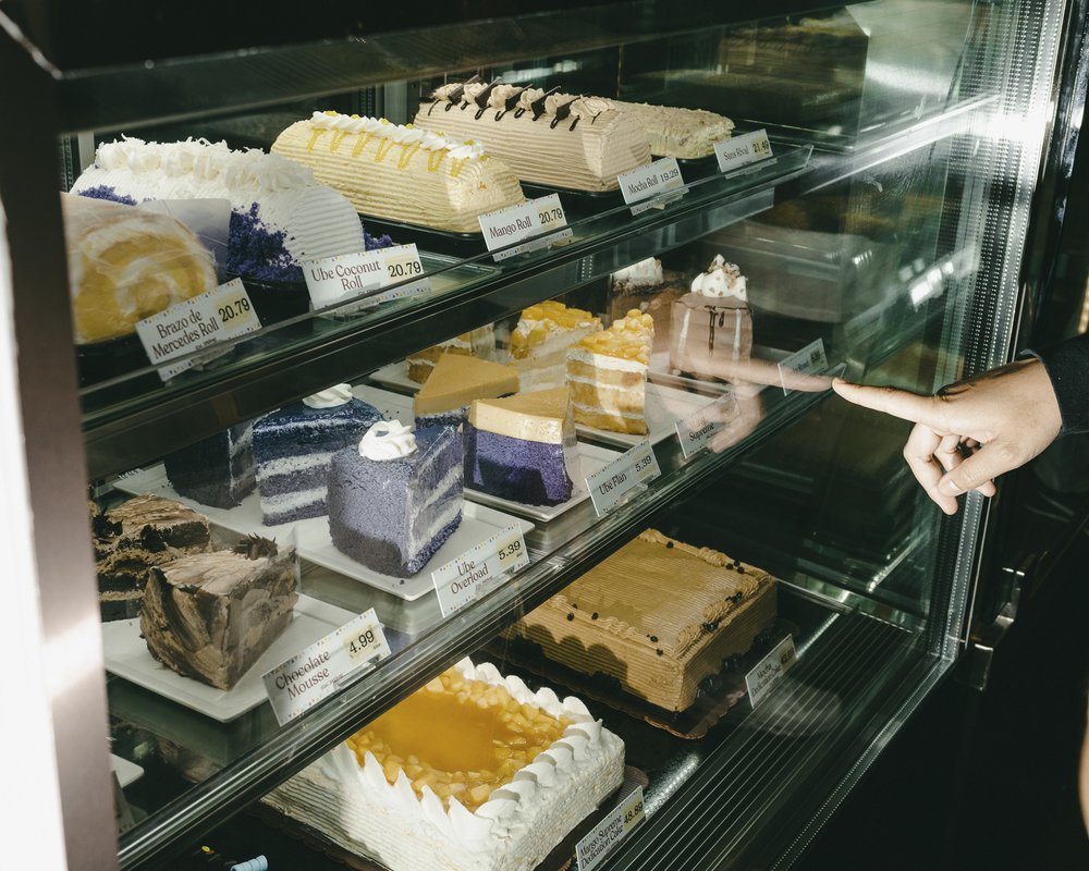 A hand points to a slice of cake. Multiple slices of cake sit inside a large glass showcase.