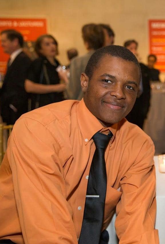 A man wearing an orange shirt smiles while sitting at a table.