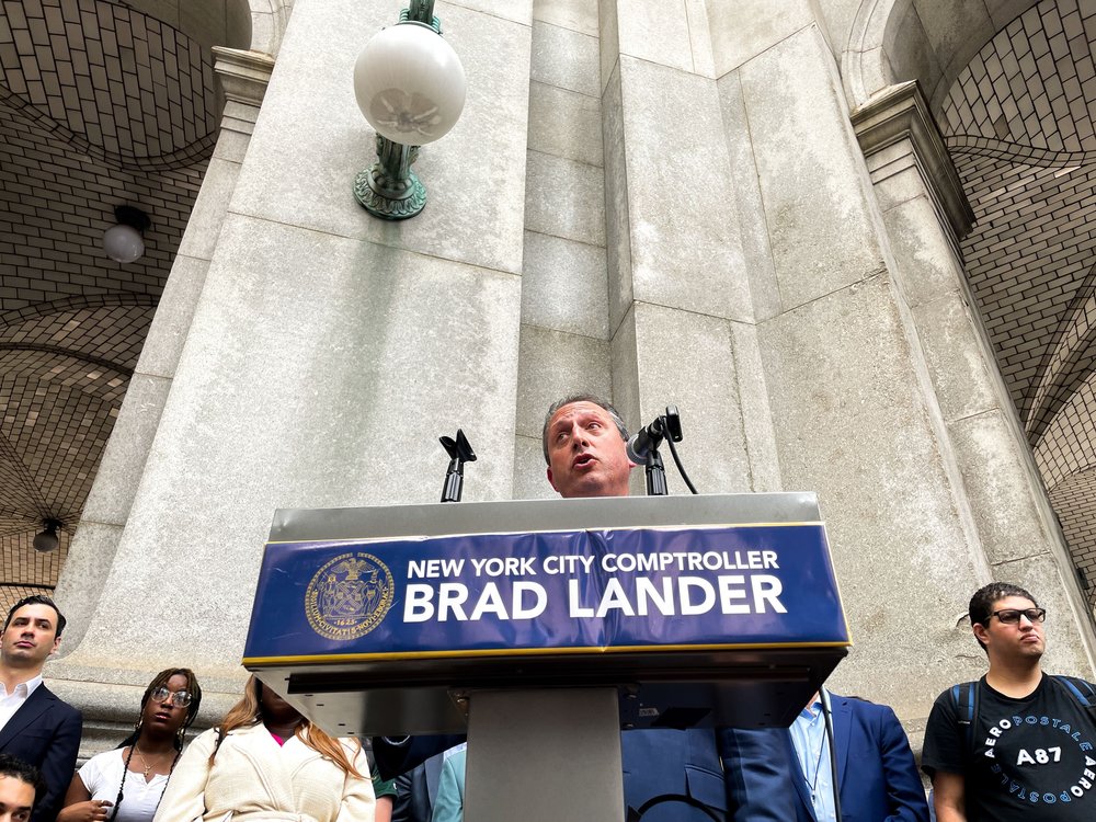 Comptroller Brad Lander speaks at a podium with a crowd behind him.