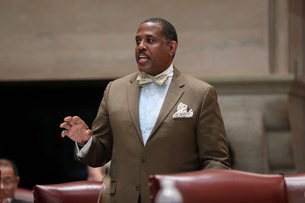 State Sen. Kevin Parker, in a tan suit and bow tie, stands in the senate chambers at the state Capitol in Albany.