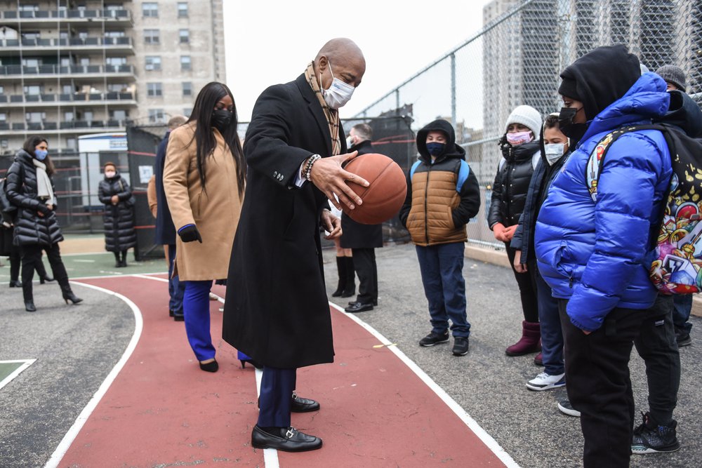 Mayor Eric ADams in a coat and mask holds a basketball in the yard, with school children watching