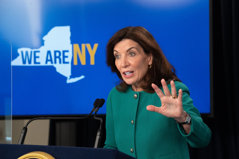 Governor Hochul in a green jacket, in front of a screen that says We Are NY