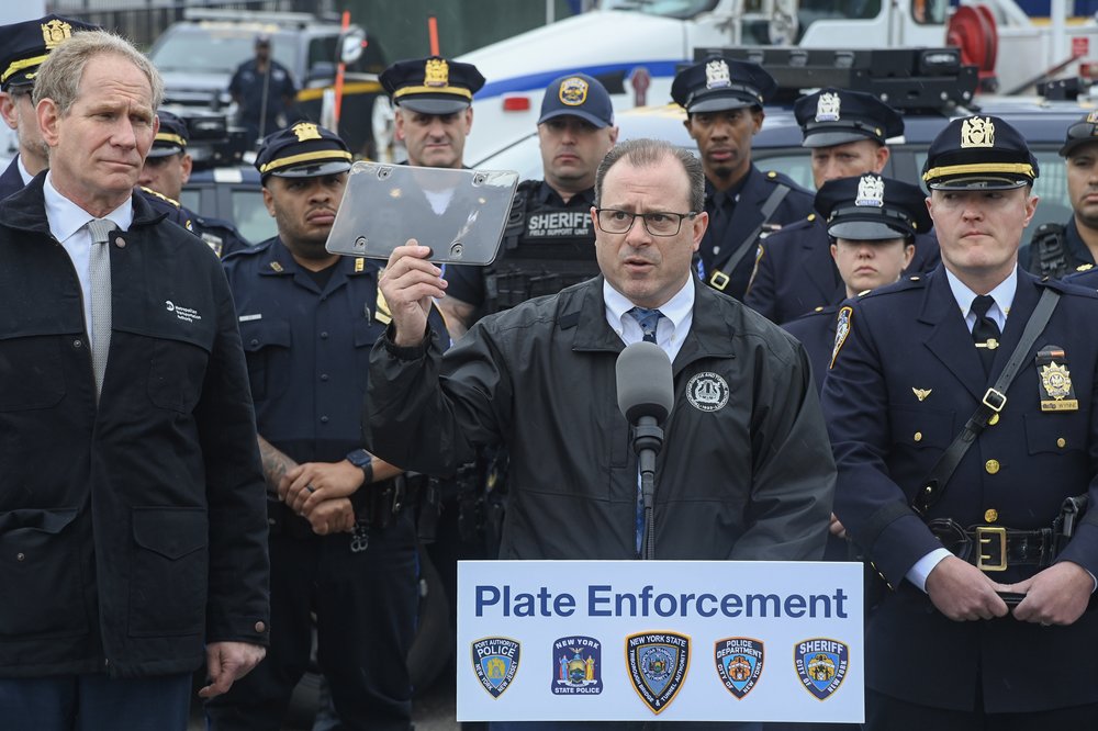 Danny DeCrescenzo in a MTA Bridge and Tunnels wind breaker, holds up a plastic screen used to obscure license plates