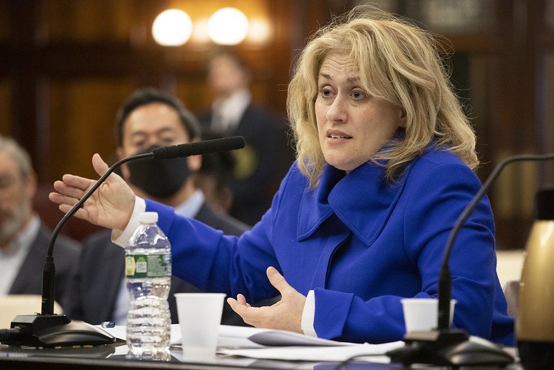Beth Rotman, in a cobalt blue coat, sits at a table with microphones and documents as she addresses the City Council's Committee on Government Operations.