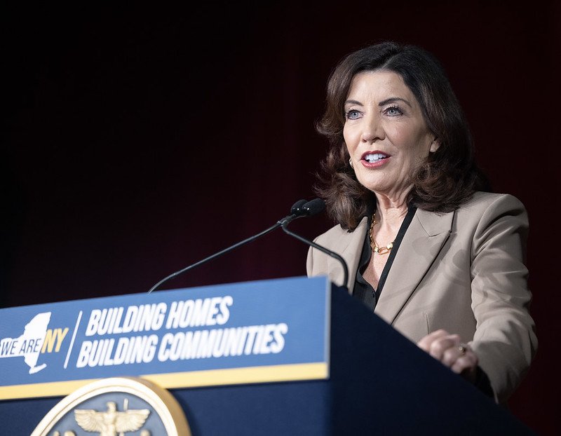 Gov. Kathy Hochul makes an announcement behind a podium that has a blue sign saying "building homes building communities."