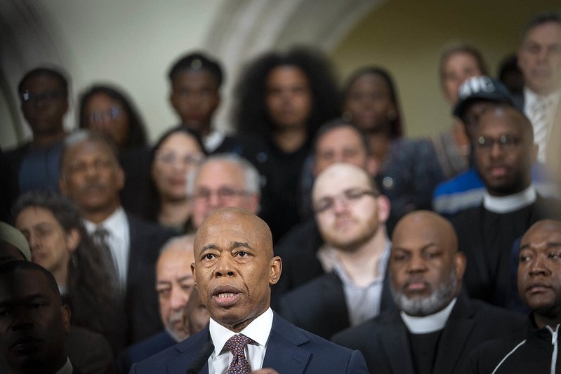 Mayor Eric Adams in a navy suit, with a crowd of people out of focus behind him.