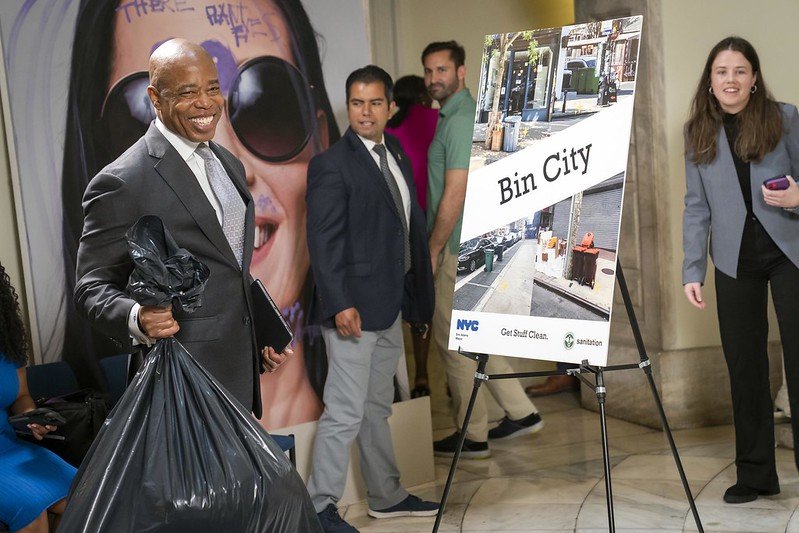 Mayor Eric Adams grins as he holds a black garbage bag, with a sign that says "bin city" behind him.