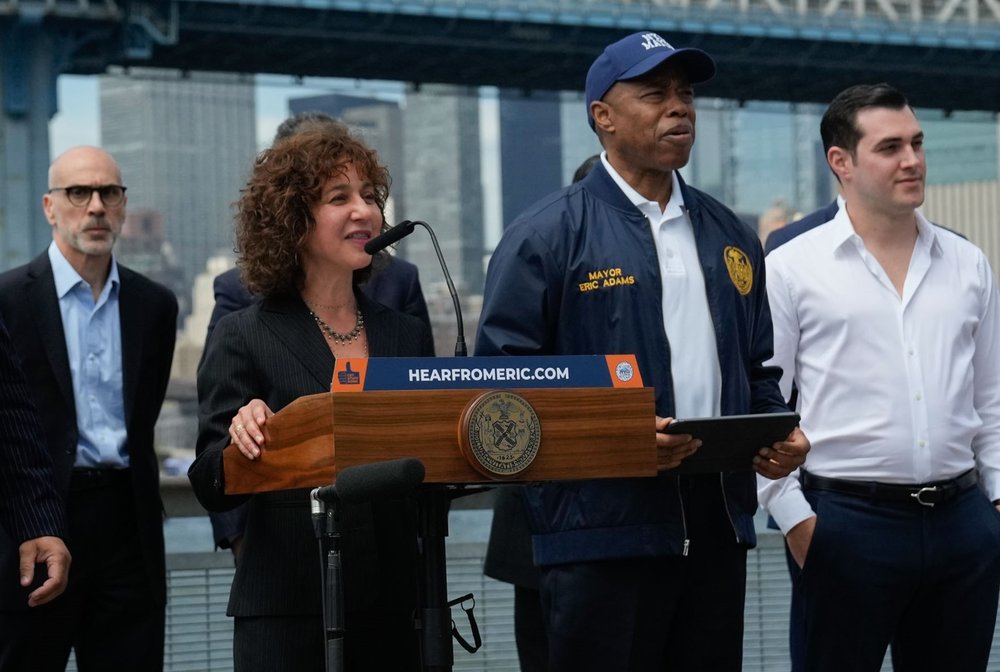 A woman at a podium stands next to Mayor Adams.
