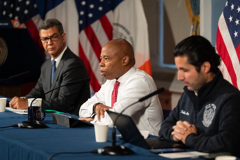 Mayor Eric Adams sits behind a table during a storm briefing, where he's flanked by two aides.