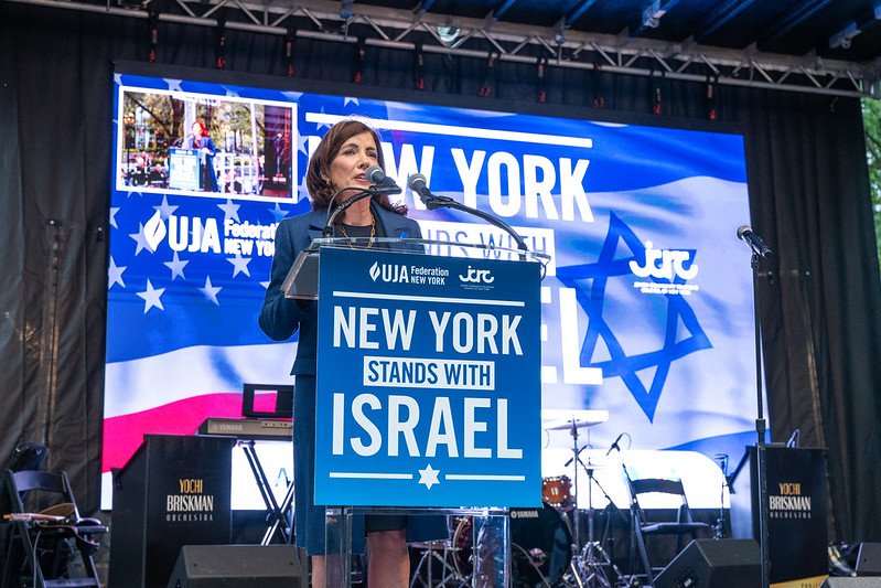 Gov. Kathy Hochul stands behind a podium with a blue and white sign saying "New York stands with Israel