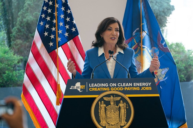 Gov. Kathy Hochul, in a sky blue suit jacket, speaks from behind a podium that reads "leading on clean energy. Creating good-paying jobs."