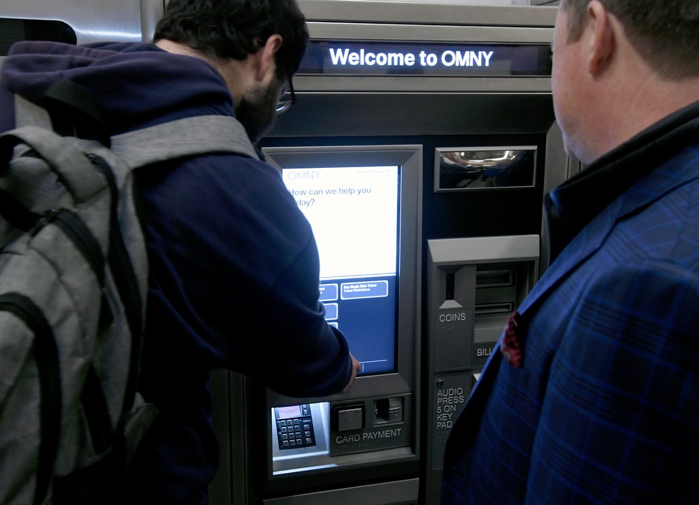 Two men stand at a vending machine with a sign reading "Welcome to OMNY."