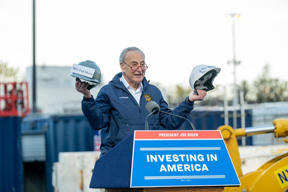 Senator Charles Schumer stands at a construction site at a podium with a sign reading "investing in America."
