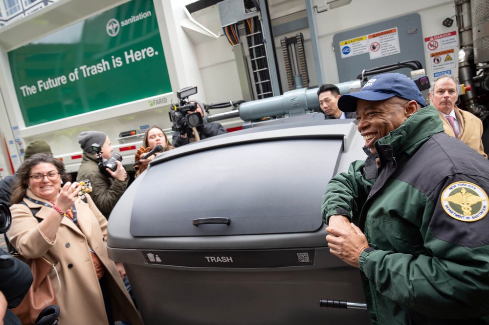 Mayor Eric Adams smiles next to a trash container, while journalist Katie Honan smiles while taking a photo with her smartphone.