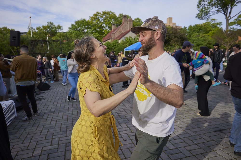 Two people dance in the street, one wearing a yellow dress.
