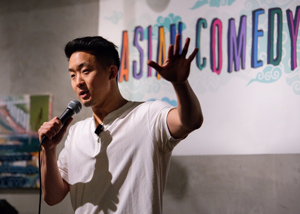 A comedian stands in front of a microphone on a stage.