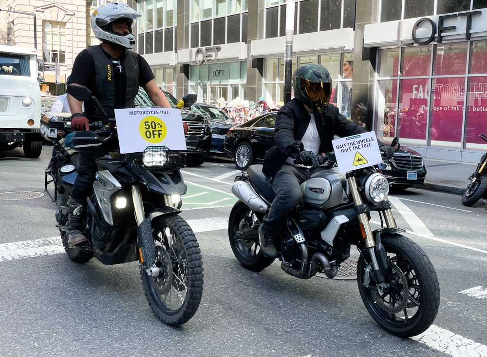 Two people riding black motorcycles with signs saying "half the wheels, half the tolls."