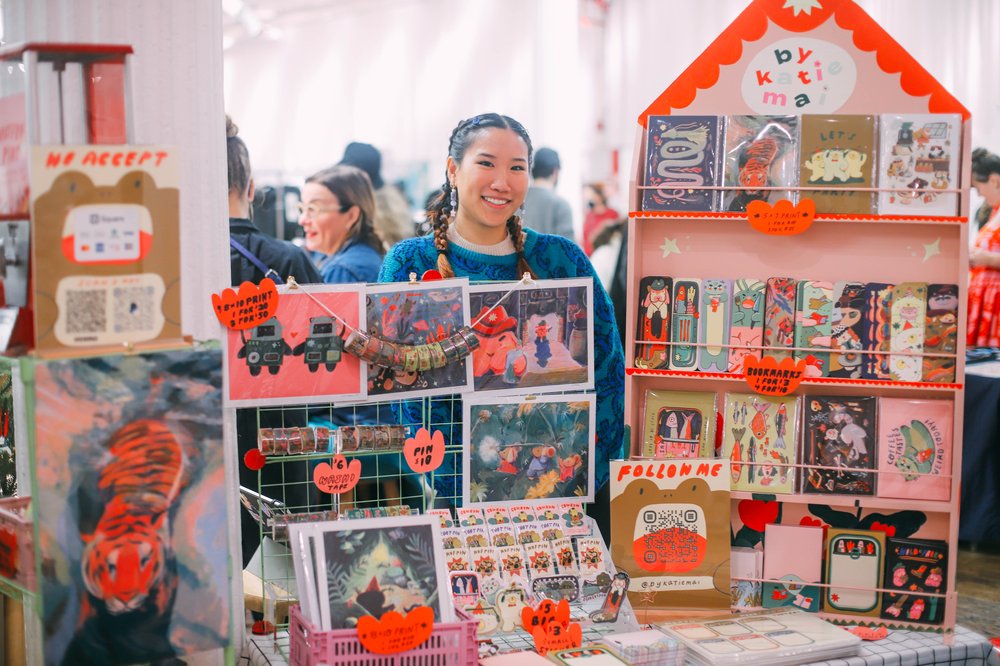 A woman stands behind crafts she's selling.