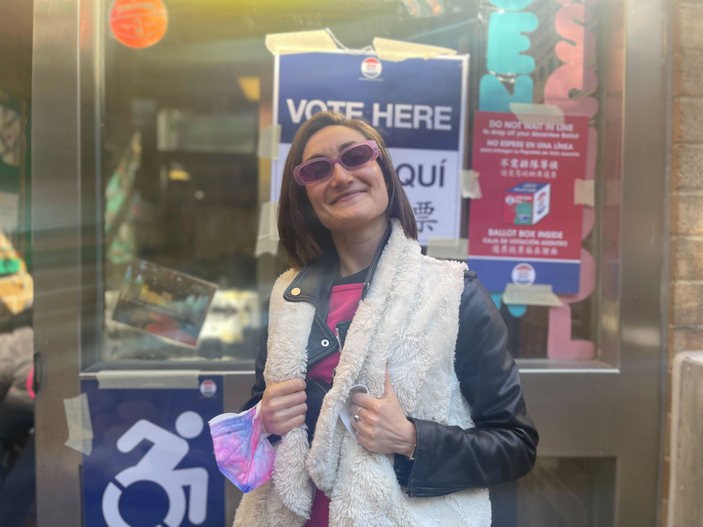 A smiling woman in a vest and sunglasses, standing in front of a polling location.
