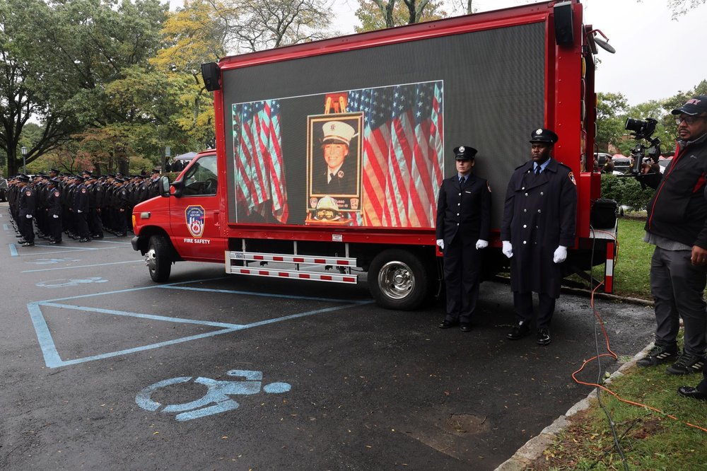 Two men stand in front of a fire truck with a screen showing a woman's picture.