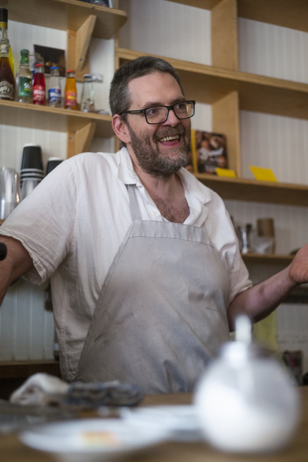 A man standing in a kitchen smiling wearing a white shirt and tan apron