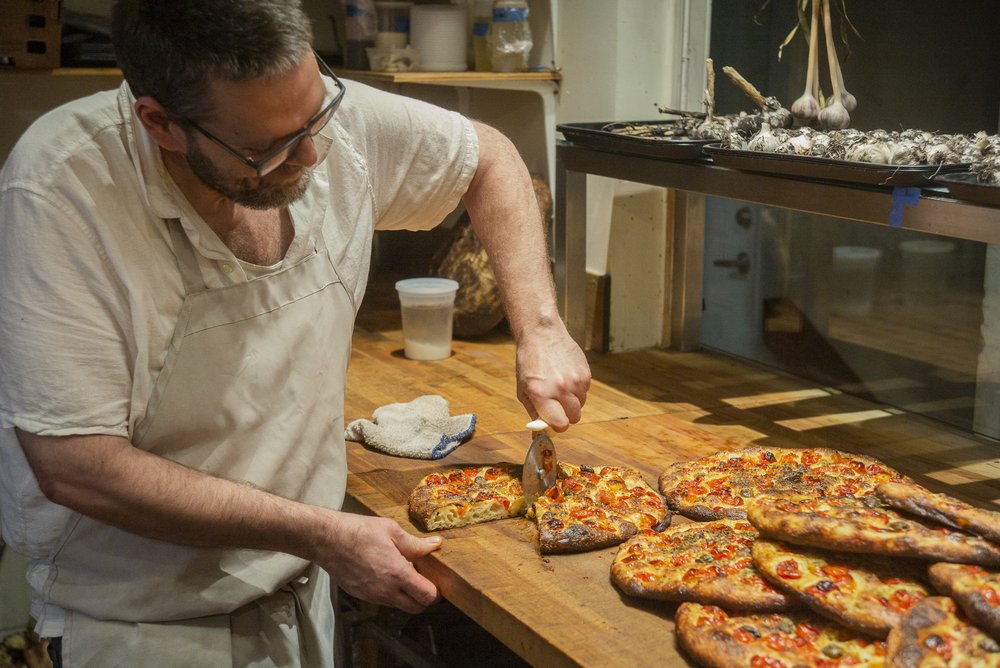 A man in a kitchen wearing a white shirt and tan apron cutting a pizza pie with a round pizza cutter