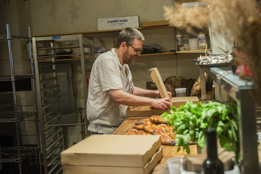 a man in a kitchen closing the lid to a pizza box