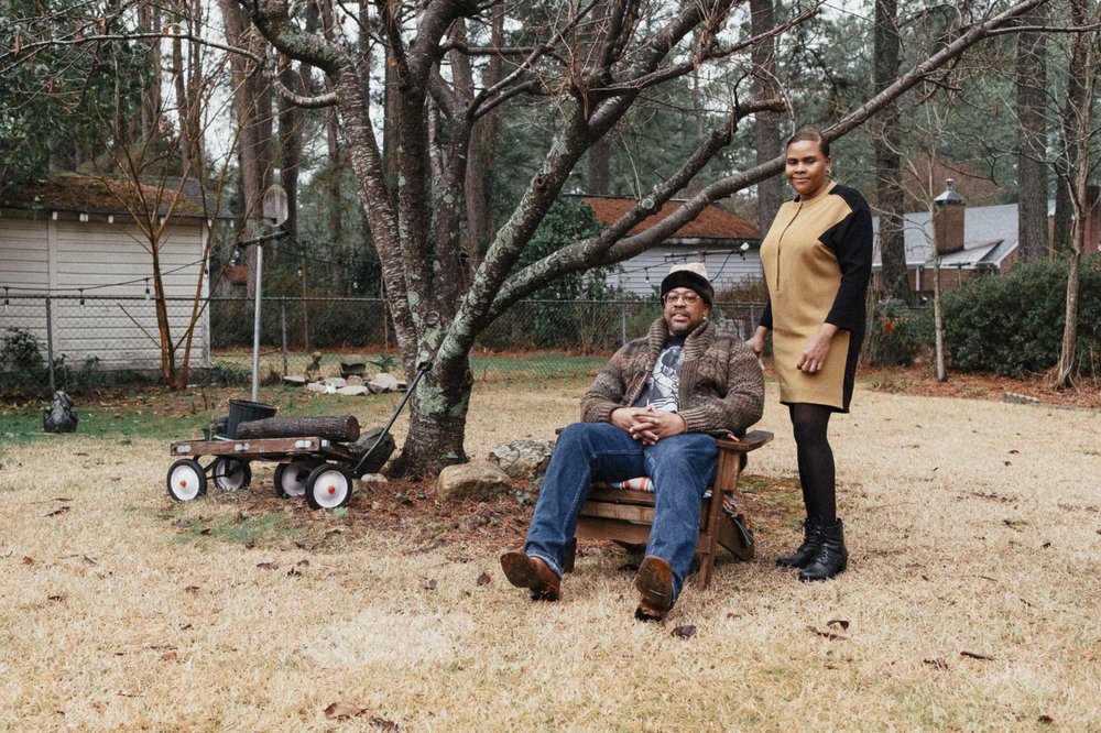 A couple photographed in their yard, beneath a tree, firewood to their left.