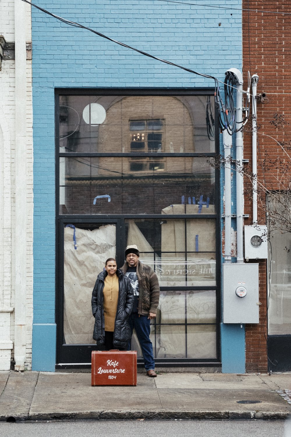 A couple stands in front of a light blue building that has brown paper in its windows, as it is under construction.