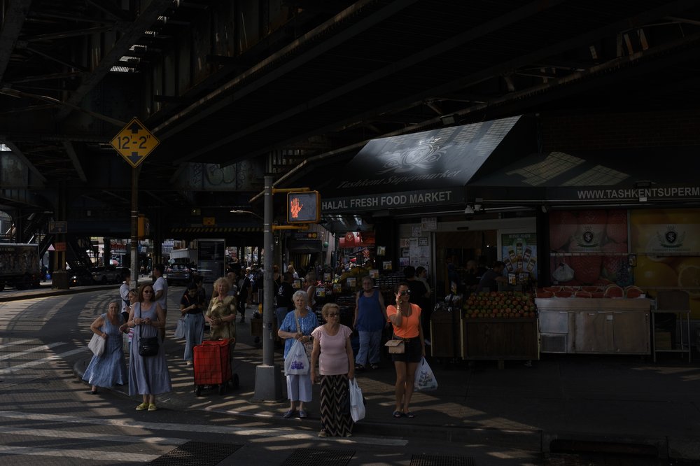 A crowd standing in front of a corner market waits to safely cross the street. The market, per the awning, is called Tashkent Supermarket.