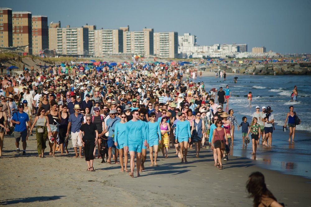 A small group of costumed dancers leading a large crowd along a beach