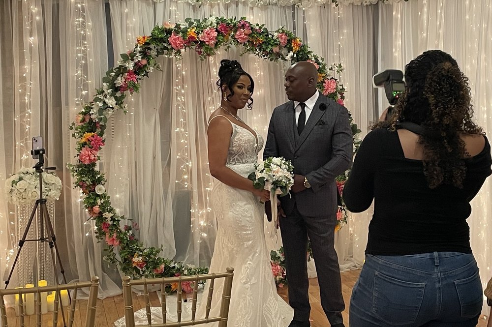 A bride and groom stand near a floral wreath for their wedding.