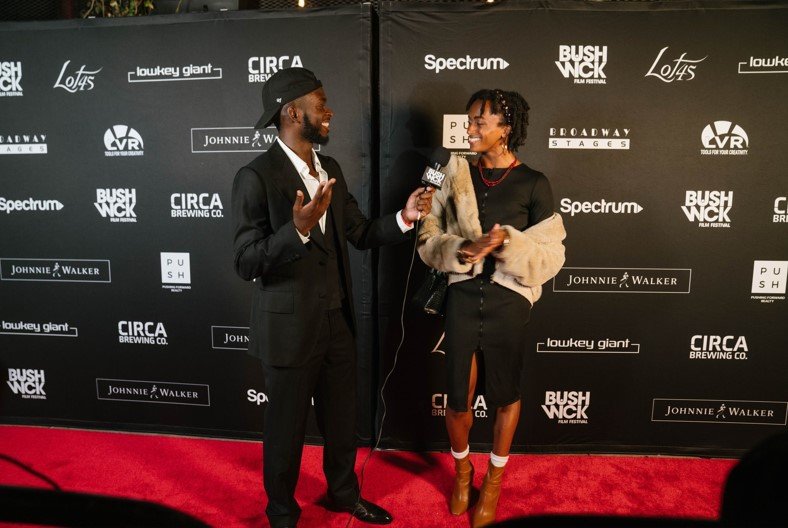 Two people stand and talk on a red carpet.