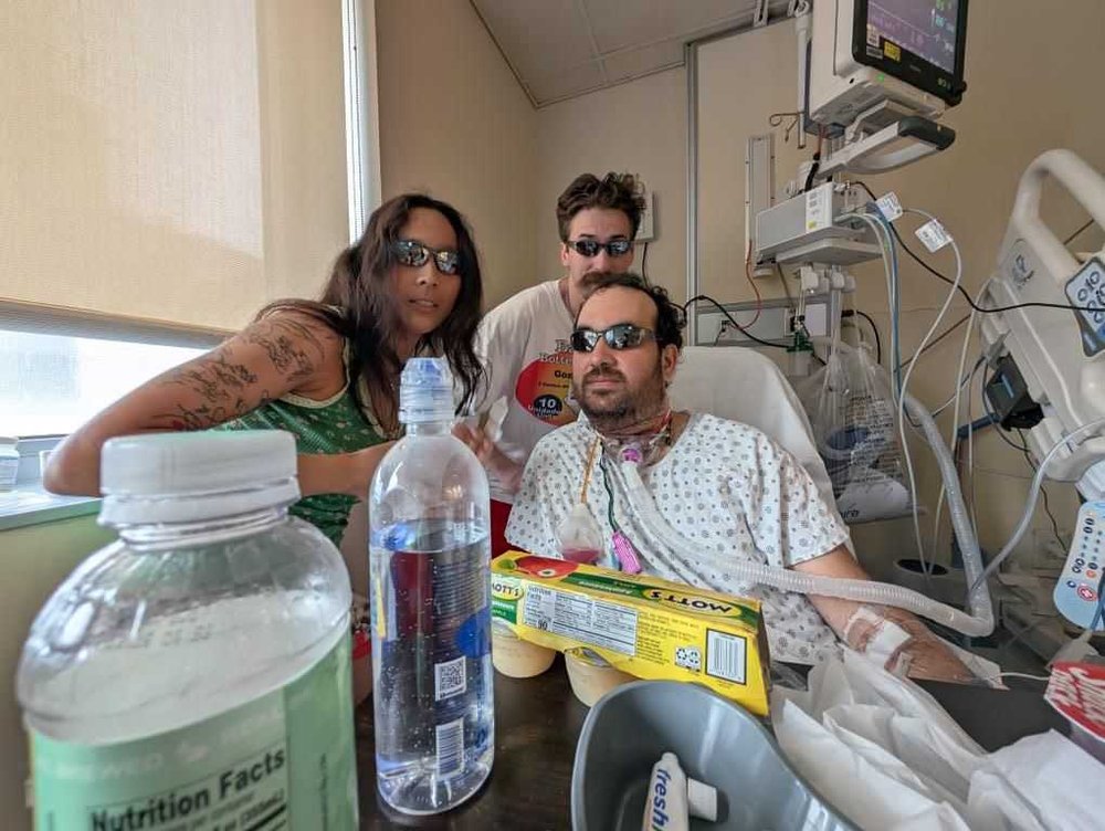 A man in a neck injury, wearing sunglasses in a hospital bed and surrounded by two friends.