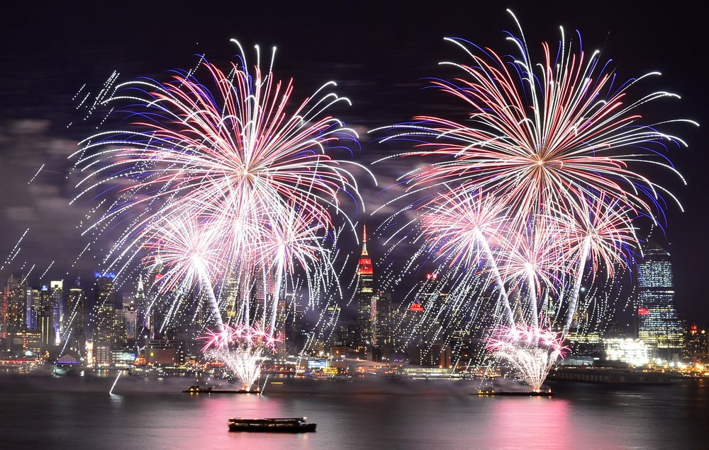 Fireworks explode over the water in NYC with a view of the Empire State Building in the background.