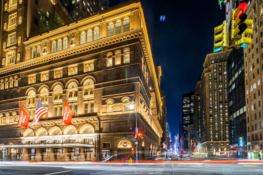 The exterior of a building, Carnegie Hall, in midtown Manhattan, with flags hanging overhead.