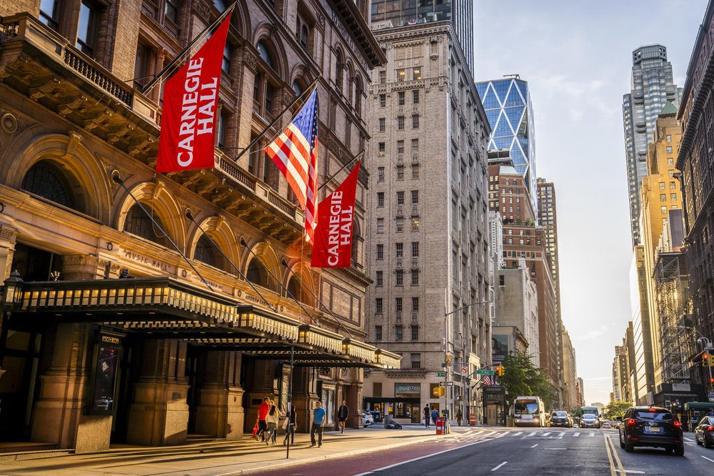 The exterior of a building, Carnegie Hall, in midtown Manhattan, with flags hanging overhead.